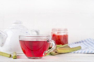 Organic natural rhubarb tea, sweet herbal plant based pink and green rhubarb hot tea drink on teapot and glass cup, on white wooden table with fresh rhubarb copy space