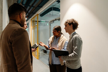 A young White man with a notebook and an Indian adult woman are talking with a white-skinned man with a tablet and a dark-skinned man while standing in an office corridor