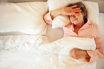 An elderly woman in pajamas is lying on a pillow in bed. Mature lady stretches and wakes up at home...