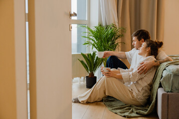 A young White man and woman sit on the bedroom floor next to the bed and window, holding coffee mugs and wearing sleepwear.
