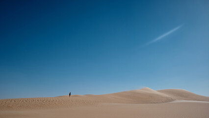 Smooth sand dunes stretching across the foreground. A single person is visible in the distance, emphasizing the scale and emptiness of the scene. Minimalistic desert landscape