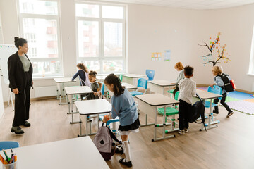 A multiracial group of school-aged children enters a brightly lit classroom with backpacks, interacting near desks. A female Black teacher in business attire watches them with a smile.