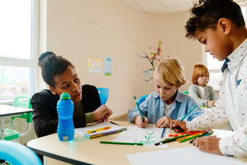 A White schoolboy and a Black schoolboy drawing together at a classroom desk under the guidance of an adult Black female teacher in a bright modern classroom decorated with colorful artwork.