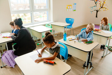 Fototapeta premium Two White school-aged boys and a Black school-aged girl are sitting at desks and drawing with pencils while their brown-skinned female teacher is talking with a school-aged boy in a classroom