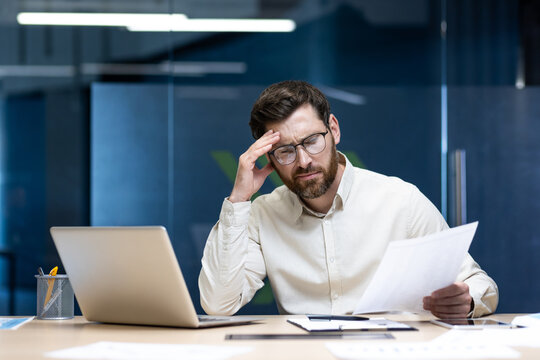Worried young businessman sitting at a desk in the office and looking at documents in frustration, holding his head - Powered by Adobe