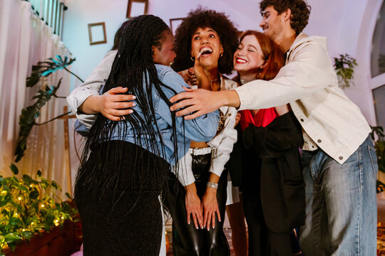 A group of diverse young friends hugs and laughs indoors during a birthday celebration, including a Black woman with braids, a White woman with red hair, and a man in a casual jacket.