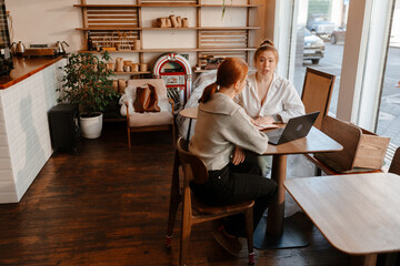 A young White female cafe owner in a white shirt discussing work matters with a young White female worker in a gray sweater at a wooden table inside a modern cafe during daytime near large windows.