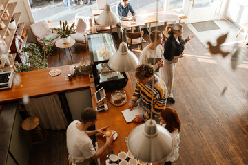 A White male barista preparing coffee behind the counter while serving a multinational group of people inside a cafe with wooden floors, wooden furniture, plants, and a window during the daytime.
