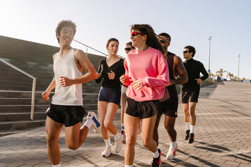 A group of diverse young athletes jogs outdoors in a sunny city, wearing sportswear and athletic shoes. The group runs on the paved sidewalk near the stairs.