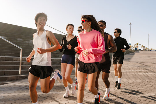 A group of diverse young athletes jogs outdoors in a sunny city, wearing sportswear and athletic shoes. The group runs on the paved sidewalk near the stairs. - Powered by Adobe