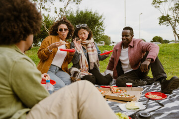 A Hispanic adult woman and an Asian woman are holding plastic plates, eating pizza, and sitting on a blanket on a grass field with a Black adult man while looking at a light brown-skinned man