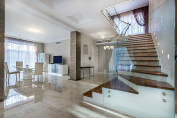 Modern interior of the living room of a luxurious country mansion. Stairs with glass railings. Beige floor tiles, light room decor.