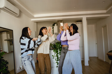 A group of multiracial adult women, including Asian, Black, and White, are dancing, laughing, and enjoying time together indoors at home. They wear casual clothing.