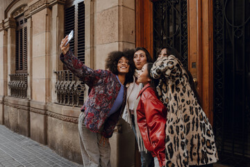 Fototapeta na wymiar A multiracial group of stylish young women takes selfies together on a city sidewalk in front of a historic urban building. They wear fashionable winter outfits.