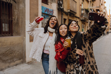 Obraz premium Three besties pose for a joyful street selfie, drinks in hand, radiating confidence and carefree energy in their stylish winter outfits. The concept of youth and women's friendship.