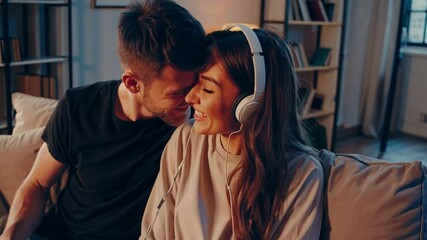 Young couple sharing headphones, listening to music and laughing together on a comfortable sofa in their living room - Powered by Adobe