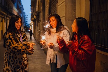 Fototapeta premium Three adult women in their 20s, including Asian and Hispanic friends, stand on a city street at night, laughing and holding sparklers. They wear winter jackets during a casual urban celebration.