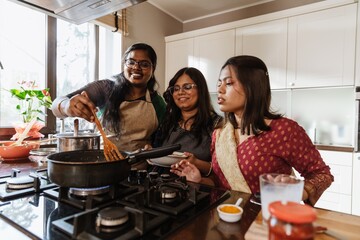 A group of young Indian women wearing aprons, cooking together in a modern kitchen, stirring a pan, while one woman holds a plate and another adds ingredients, smiling and enjoying the moment.