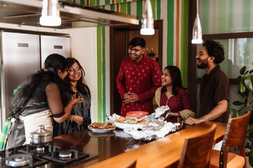 A group of young Indian people wearing traditional outfits gathered around a kitchen island indoors, laughing and preparing dinner together, plates of Indian food on the counter in a modern home.