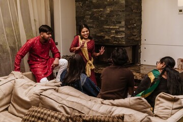 Five South Asian adults, including men and women in traditional clothing, sit and talk in a modern living room. One woman stands and gestures while the others listen and smile.