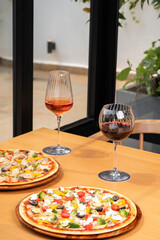 Grilled vegetable pizza and cajun-spiced chicken pizza served on a brown table with two cocktails in the background, framed by blurred windows and plants.