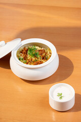 Fragrant vegetable biryani with saffron, herbs, served in a white pot with lid, paired with curd in a bowl—captured close-up under soft sunlight on a brown table.      