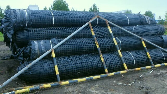 Black plastic mesh rolls wrapped plastic film, supported metal structure, ready use in road construction stabilize ground, prevent erosion. Plastic mesh rolls for road construction stabilizing ground