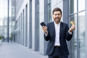 A frustrated businessman, holding a smartphone and a credit card outside an office building, appears to be in a stressful situation.