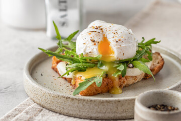 Toasted bread with poached egg, cheese and arugula on a plate on white kitchen background. Healthy breakfast, diet food