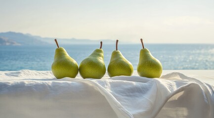 Four green pears rest on white fabric, draped over a surface overlooking a calm sea and distant mountains under a bright sky