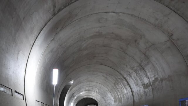 Smooth concrete tunnel walls illuminated by vertical work lights stretch into the distance subtle reflections and poster boards hint at preparation for presentation or inspection inside construction z