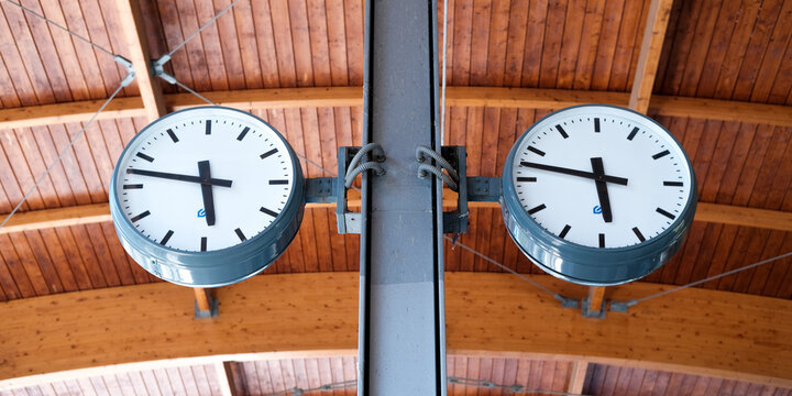 Two round station clocks with black hands and a white dial are mounted on a metal pole and show the time against a wooden plank ceiling. Railway banner with copy space