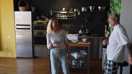 A white, middle-aged man in a white t-shirt and plaid pants and an adult woman with blond hair in casual clothing are dancing and laughing together in a cozy modern kitchen at home. - Powered by Adobe