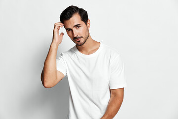 Plakat Attractive young man touching hair smiles confidently while posing in a casual white t-shirt against plain light background. Modern lifestyle concept.