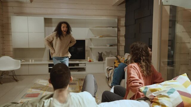 Four adults, including a White man and a multiracial group, play a guessing game in a modern living room. One woman performs in front of the others, reacting and pointing indoors.