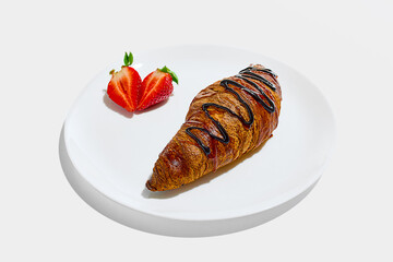 White plate with chocolate-drizzled croissant next to sliced strawberry halves arranged neatly against white studio background. Concept of traditional, takeaway, fast food, catering, nutrition.