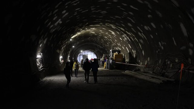 Underground tunnel construction scene with workers in helmets walking under patchy lights beside heavy machinery gritty textures echo raw progress and teamwork deep beneath the surface