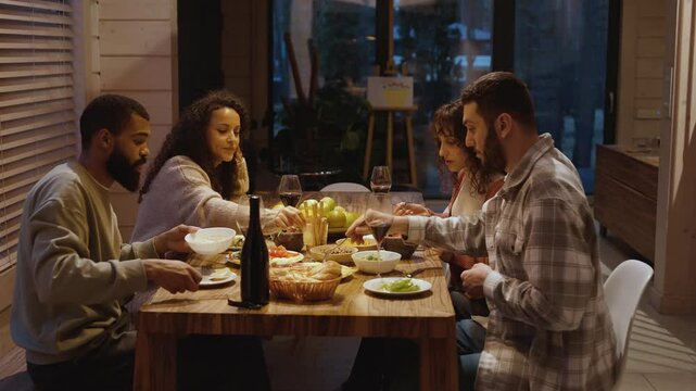 A black adult man, a White male, and two multiracial adult women sit together at a wooden table sharing a meal with wine, fruits, and appetizers in a modern home interior.