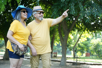 Happy indian senior couple wearing hat having eyewear sunglasses at park. Senior people enjoying retirement life. Senior Couple Enjoying Picnic Together having fun and pointing fingers aside way