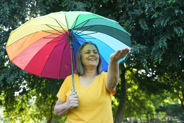 Monsoon Bonanza Concept. Portrait of Indian Senior woman under rainbow umbrella and walking outdoor...