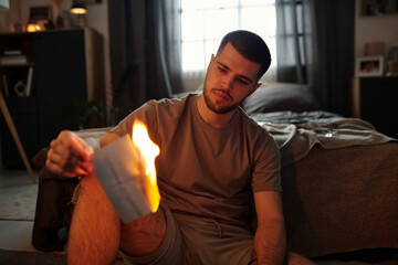 Young man sitting in cozy bedroom setting and gazing at burning envelope in his hand. Room features...
