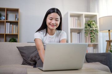 Smiling asian teenage girl using a laptop on the couch in a cozy living room.