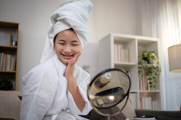 Cheerful teenage girl in white bathrobe enjoying self-care time in living room, applying skincare serum.