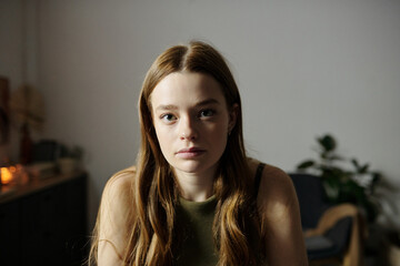 Portrait of young woman with long brown hair and green top sitting in room with dim lighting and...