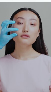 face portrait of healthy attractive chinese woman standing on grey background. Cosmetologist hands checking skin njecting hyaluronic acid into cheecks. beauty center.