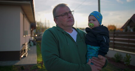 Father holding baby with pacifier in backyard, gently adjusting child’s outfit, showcasing...
