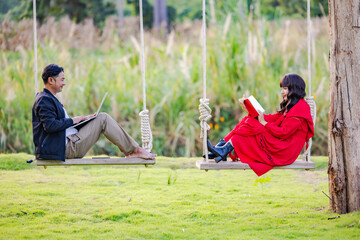 Happy couple enjoying in travel using laptop watching pictures on the laptop and reading book while sitting on the swing at beautiful blooming reeds grass flower blowing from wind.