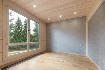 A spacious room decorated with light wood and gray textured plaster. Panoramic window .with a view of the summer landscape. Unfurnished.