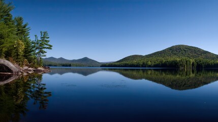 Obraz premium Panoramic view of a lake with a beautiful blue sky and forest in the background, where the water perfectly reflects the scenic landscape.