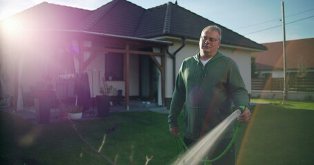 Man watering garden with hose in suburban backyard, standing near house with patio and lawn, serene...